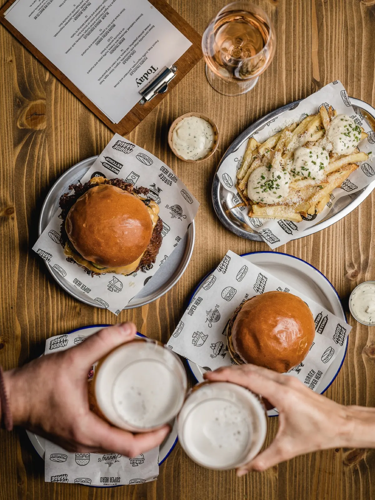 Mesa con dos burgers, patatas con salsa y cervezas brindando
