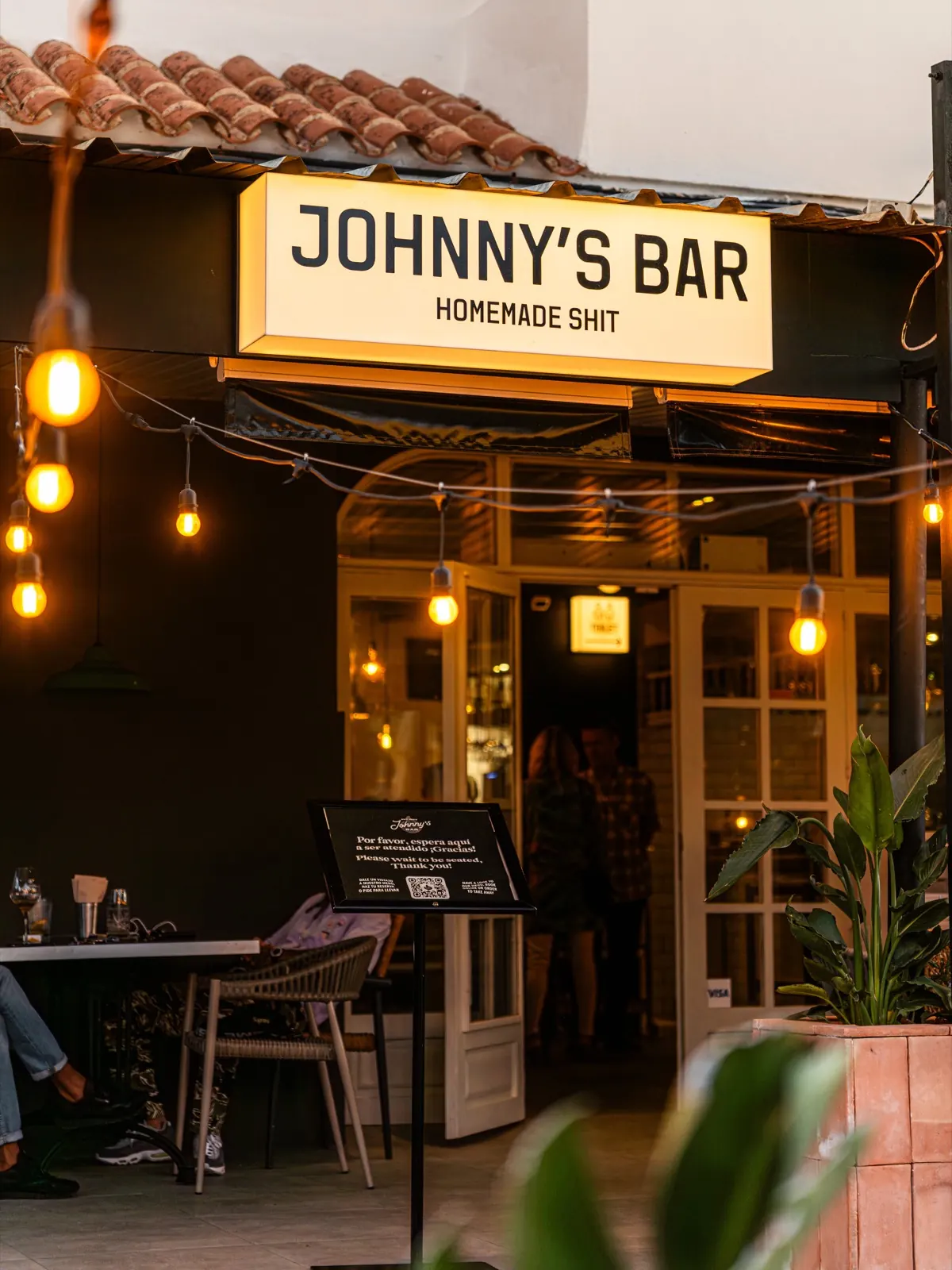 Entrada de Johnny's Bar con el cartel Homemade Shit y luces al anochecer